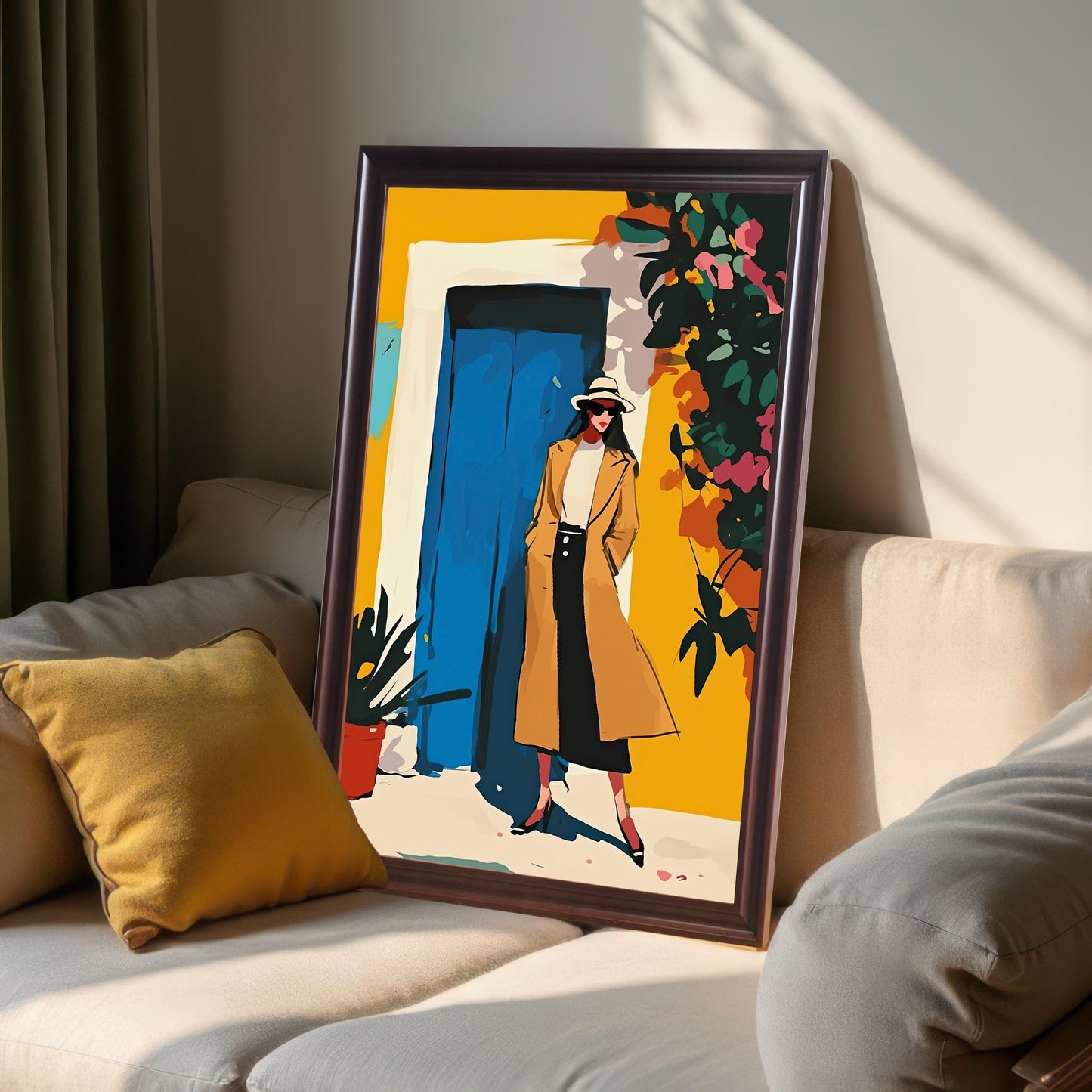 A framed art print with a coffee-colored frame is leaning against a wall on a cream-colored sofa. The art shows a woman in a tan coat standing in front of a yellow wall with a blue door. Sunlight casts a shadow across the print and sofa.