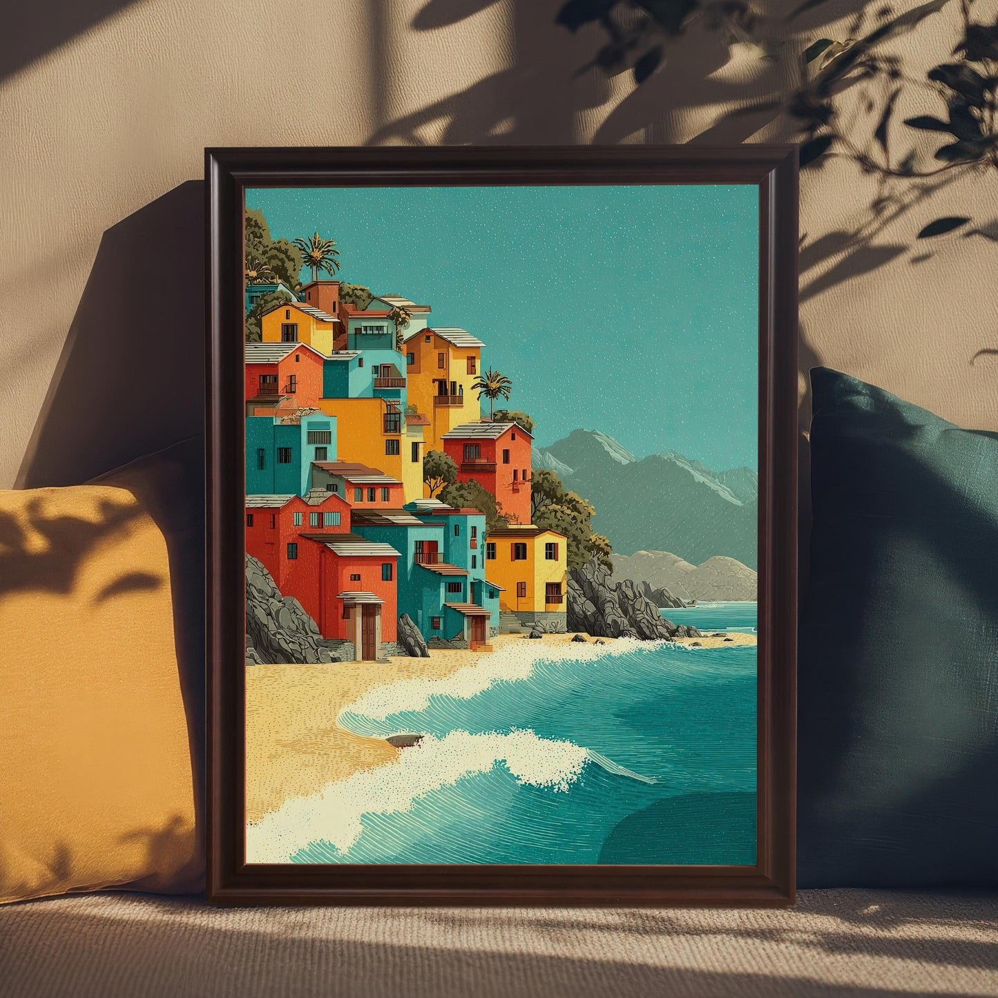 A framed art print sits on a couch, propped up against a yellow and a green pillow. The print shows colorful houses on a hill next to a beach and ocean.