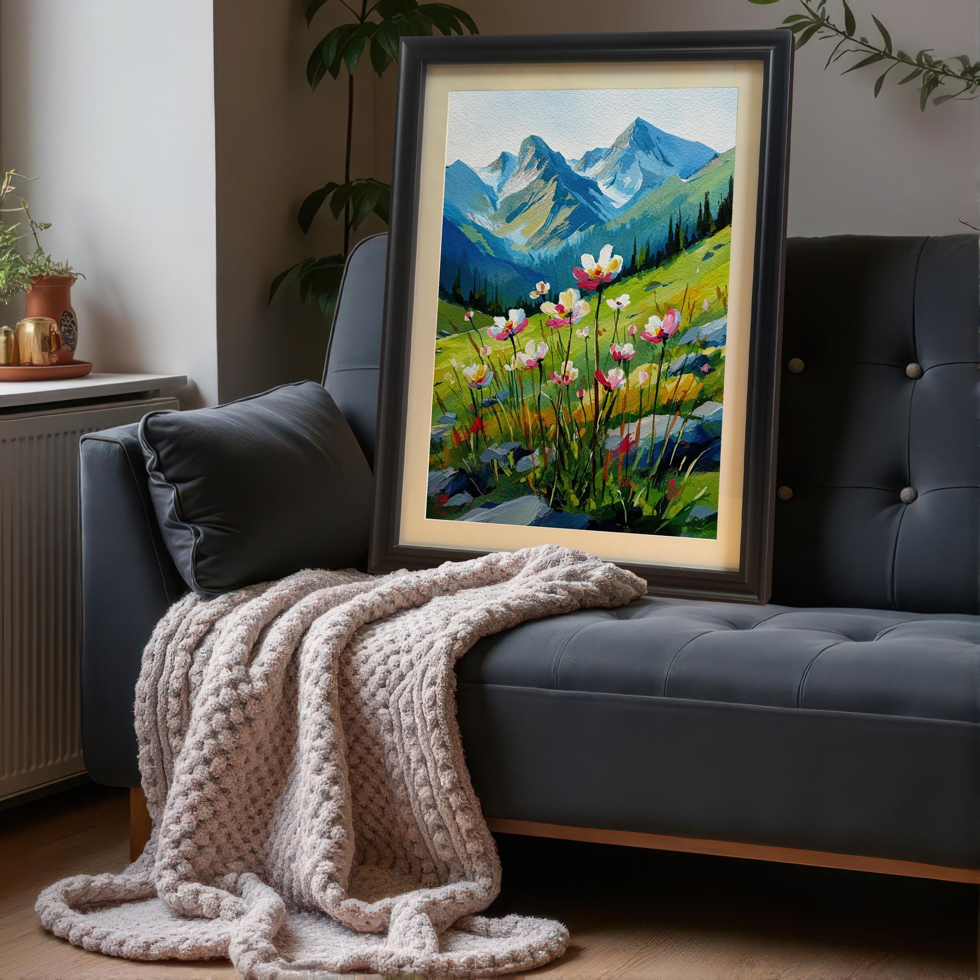 A framed landscape painting is propped on a dark gray couch with a light-colored knitted blanket draped over it. The painting depicts a field of flowers with mountains in the background and has a black frame and a mat.