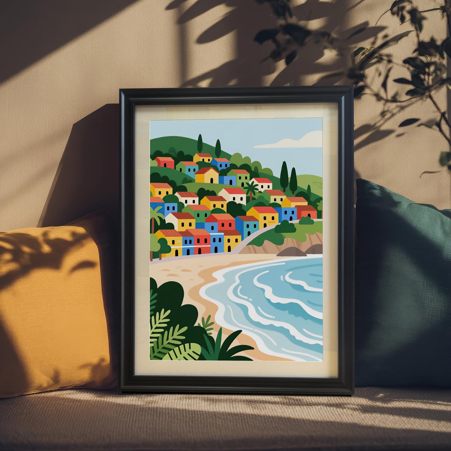 A framed painting of a village with colorful houses on a hill overlooking a beach. The painting is in a black frame and is leaning against a wall, propped up by two pillows on a couch.