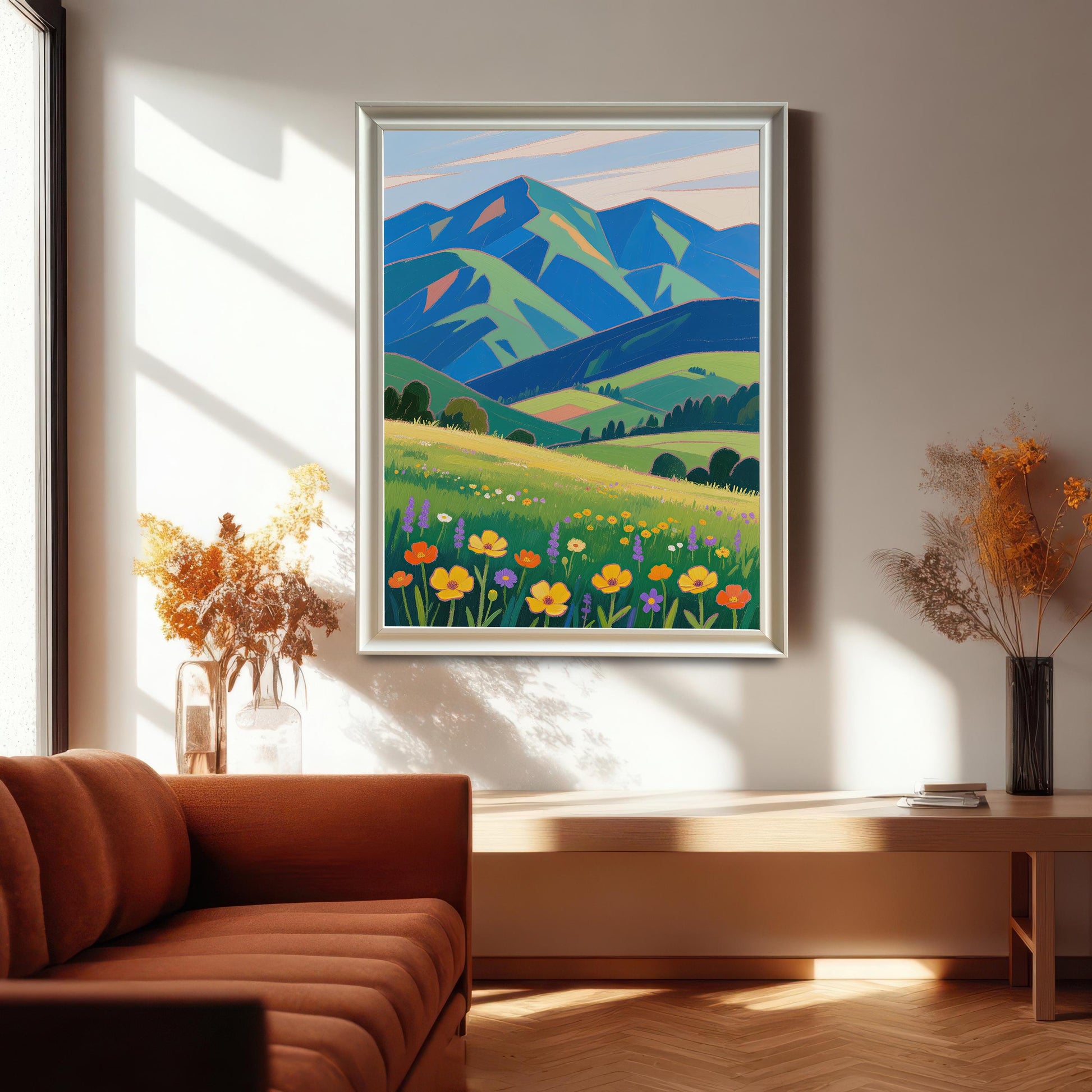 A framed painting of a meadow with colorful flowers and mountains hanging on a wall above a wooden table. A brown couch is visible in the foreground.