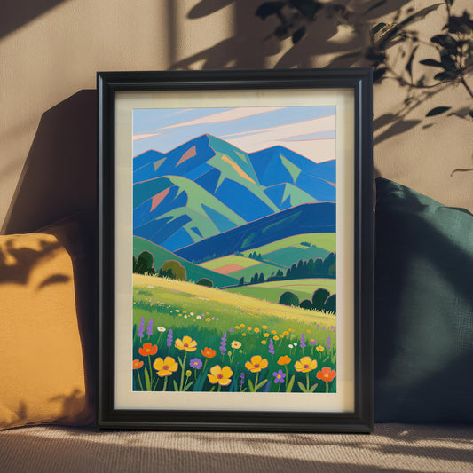 A framed painting of a meadow with colorful flowers and green and blue mountains in the background. The painting is in a black frame and is propped up on a couch between two pillows