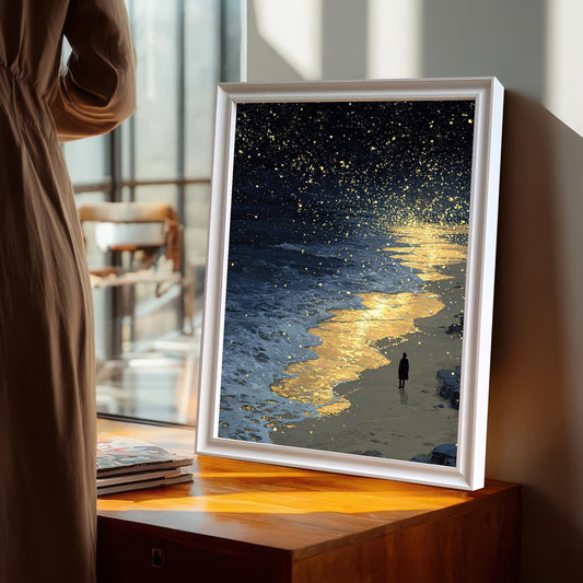 A framed painting of a dark, starry night sky over a beach. Golden light reflects on the water as a person walks along the shore. The painting is in a white frame, standing on a wooden surface next to a window.