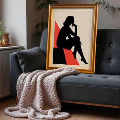 A large, gold-framed art print is propped up on a dark gray couch, resting against a plush gray pillow and a knit blanket.