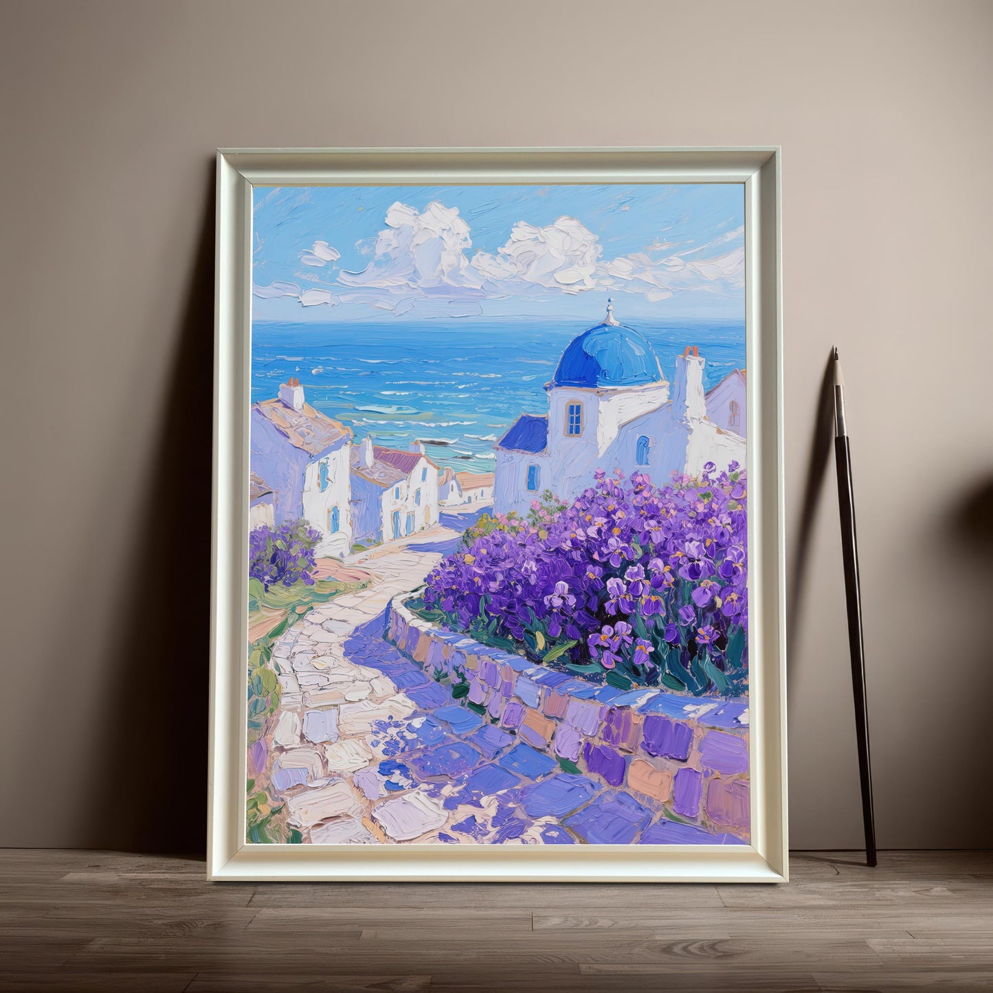A framed painting of a Greek village with a blue-domed church and purple flowers rests on the floor against a tan wall. The frame is white.