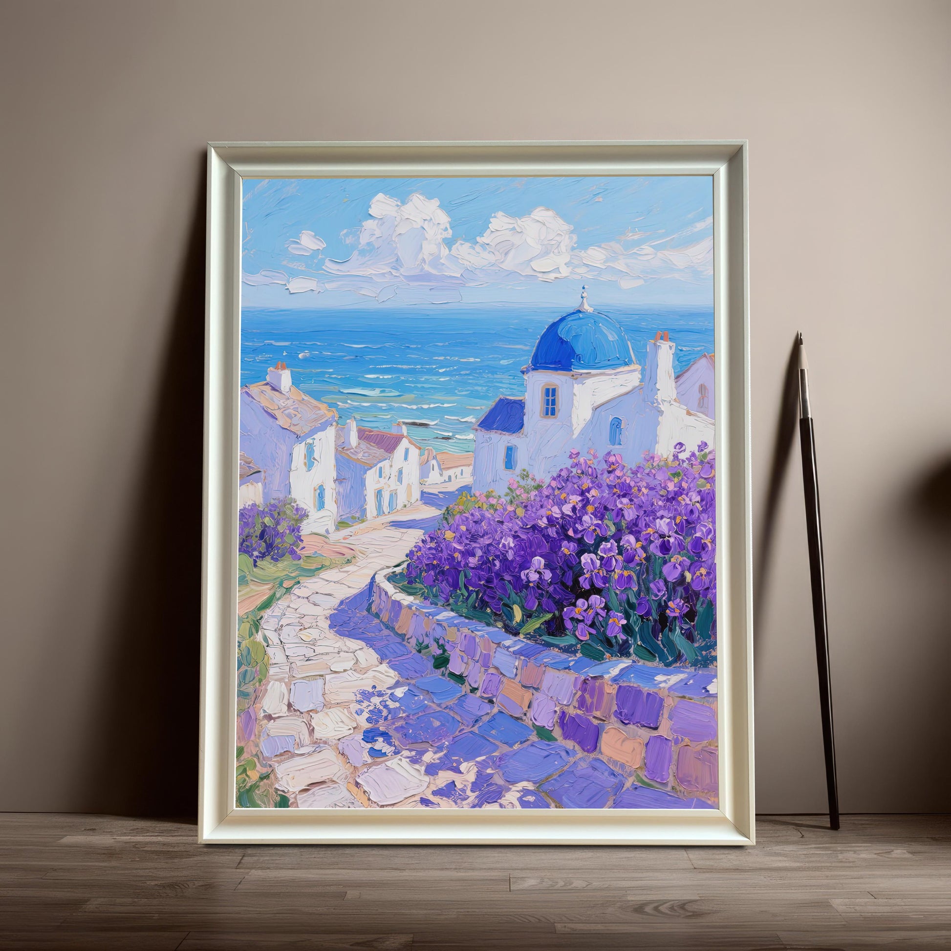 A framed painting of a Greek village with a blue-domed church and purple flowers rests on the floor against a tan wall. The frame is white.