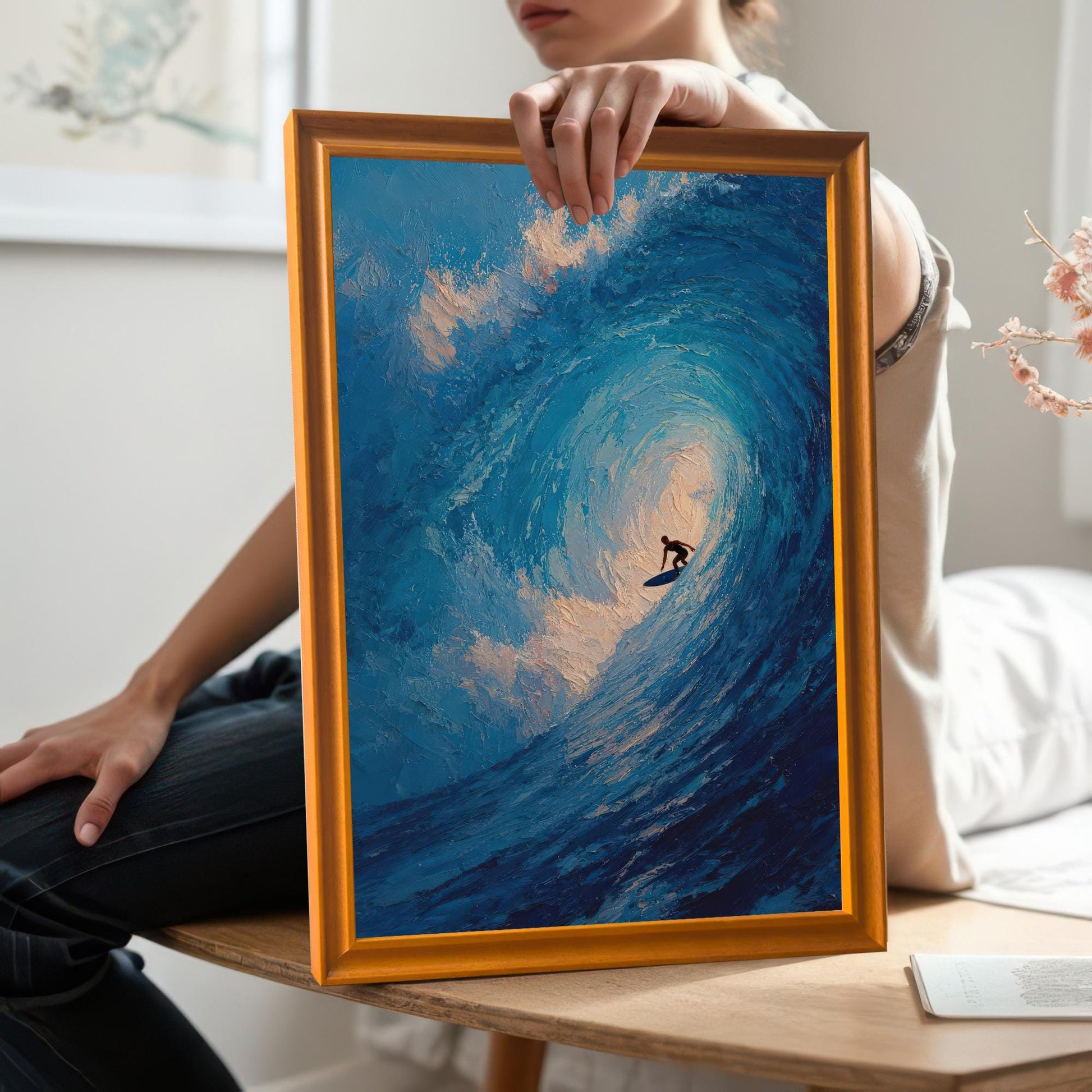 A person holding a framed print of a surfer on a wave. The frame is a light orange wood color, and the print is held up for display. The person is wearing jeans and a white t-shirt.
