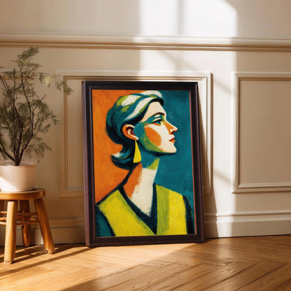 A framed print of a woman's abstract portrait. The frame is dark brown and the print is propped on a wooden floor against a white wall with ornate molding. There is a potted plant on a stool beside it.