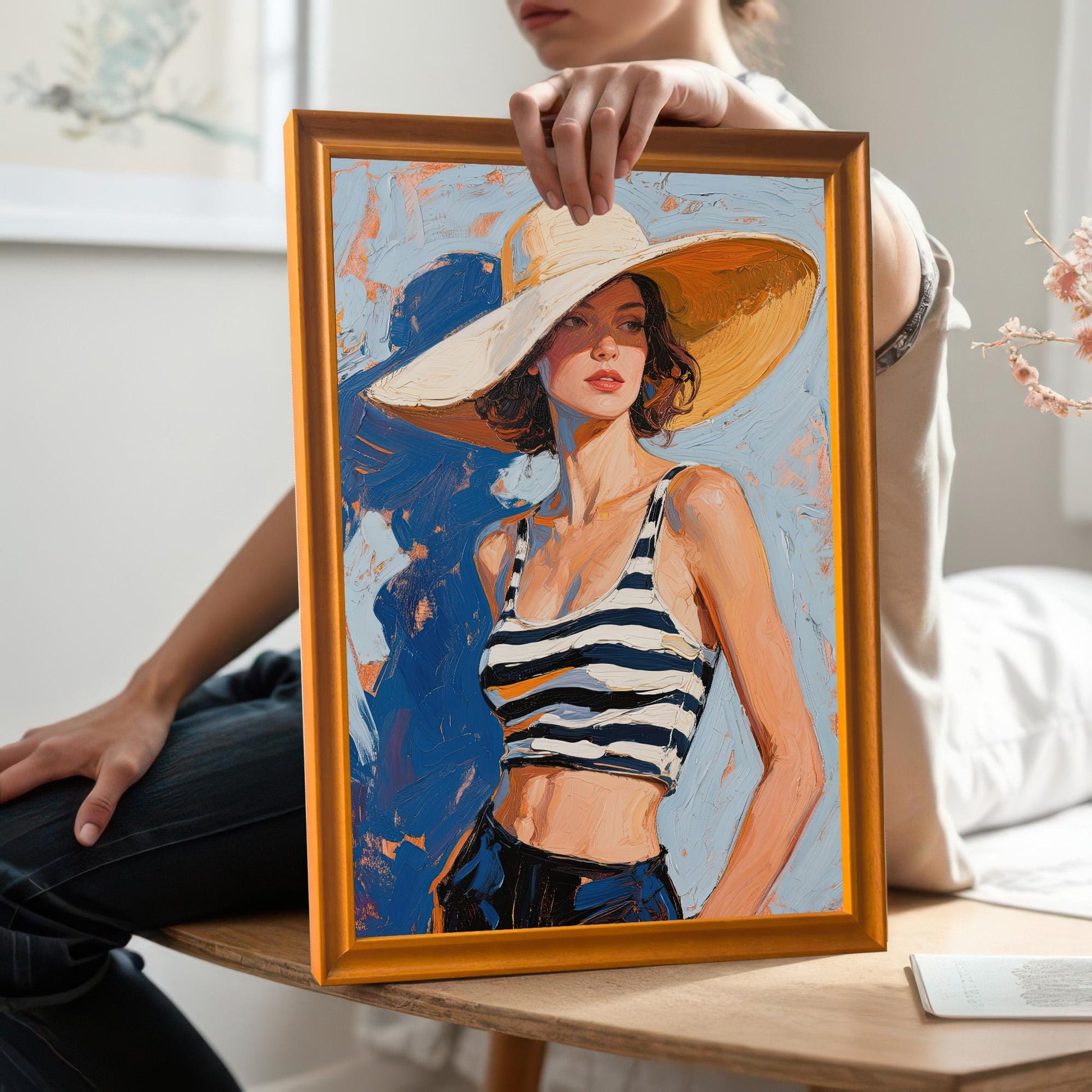 A person holds up a framed art print of a woman in a striped top and large sun hat. The frame is orange.