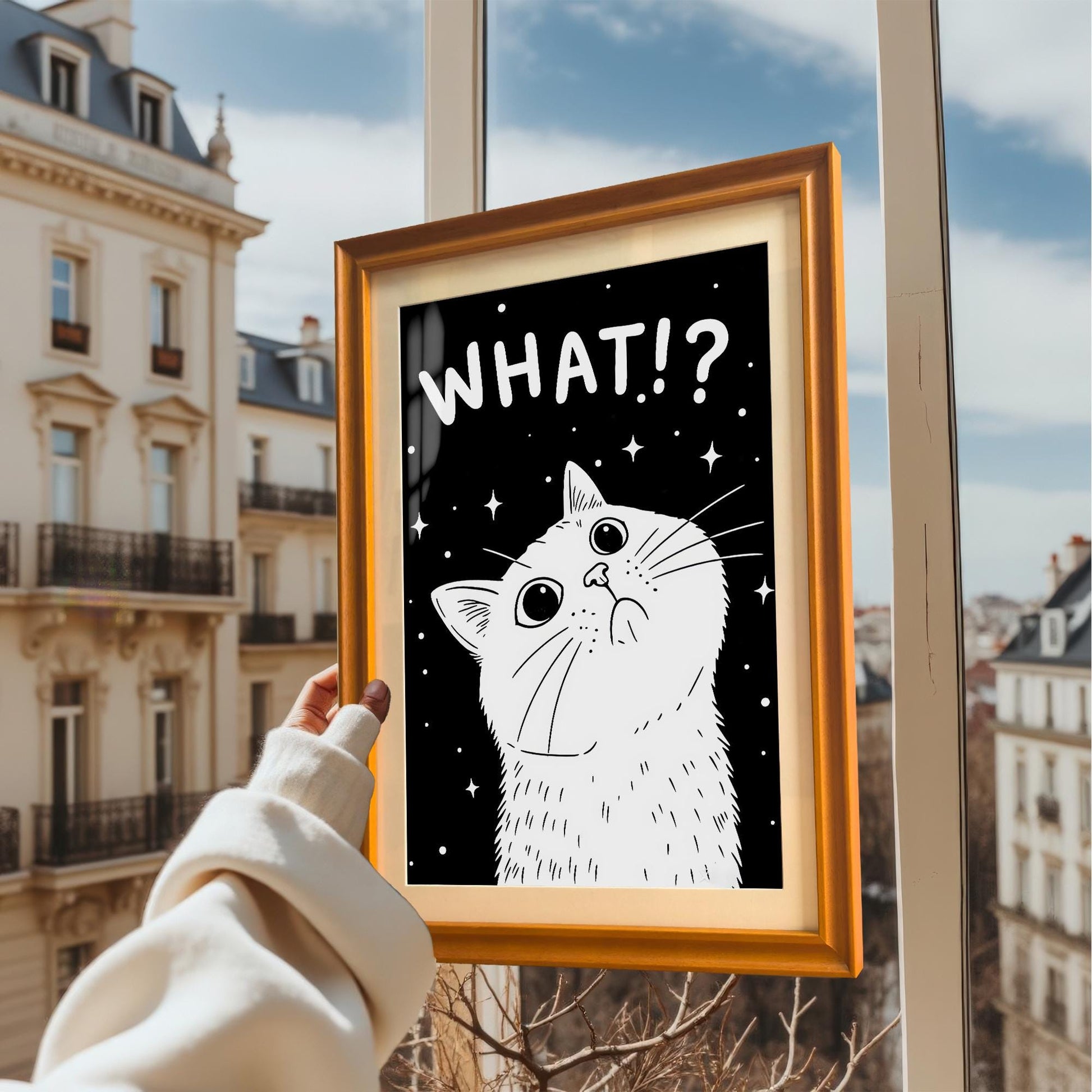 A person holds a framed art print of the black and white cat with "WHAT!?" up to a window with a view of European-style buildings and a blue sky.