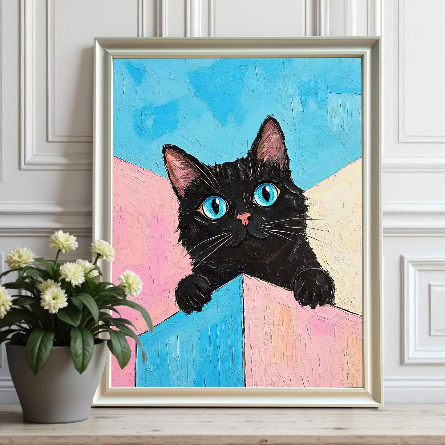 A white-framed painting of a black cat with vibrant blue eyes sits on a wooden surface next to a potted plant with white flowers.