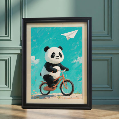 A framed print of a cute panda riding a bicycle, with a blue sky and white clouds in the background. The frame is black. The image is a full shot of the framed art leaning against a green paneled wall.