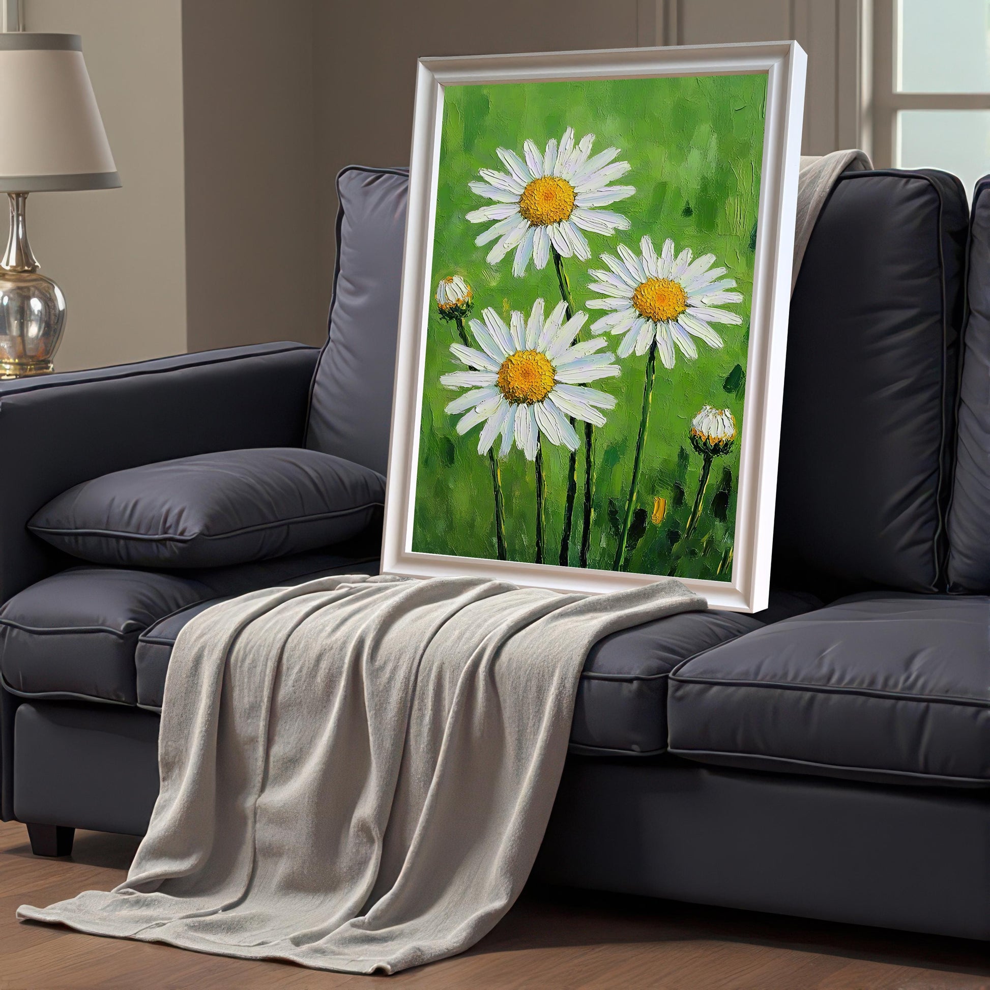 A framed daisy print with a white frame propped on a dark gray couch with a light gray blanket draped over it.