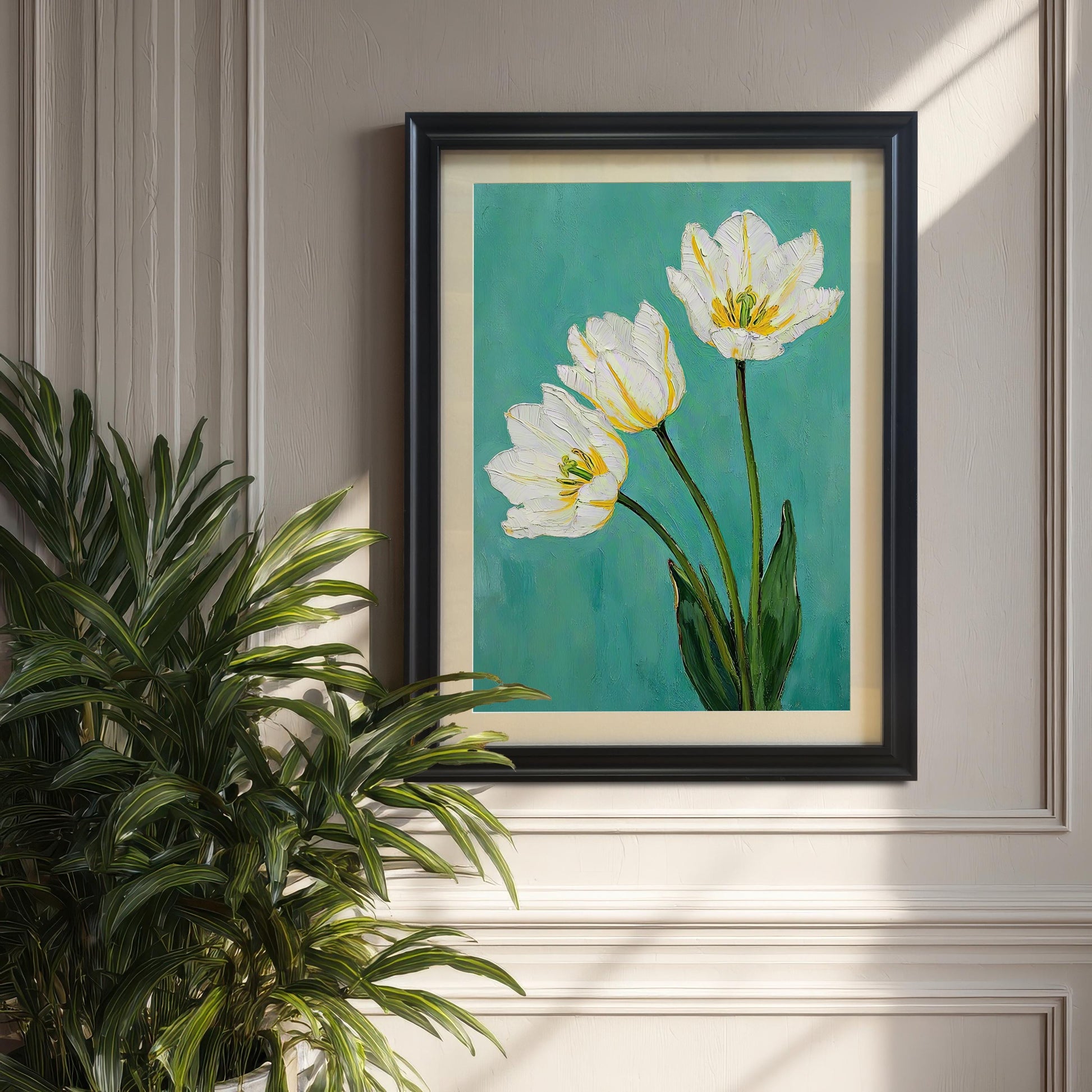 A framed art print of three white tulips with green leaves on a teal background hangs on a light gray wall. The print has a black frame and a tan mat. A large green plant is on the left.
