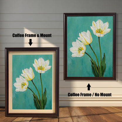A framed art print of three white tulips. One is in a coffee-colored frame with a tan mat, and the other is in the same frame without a mat. The art is against a white wooden wall.