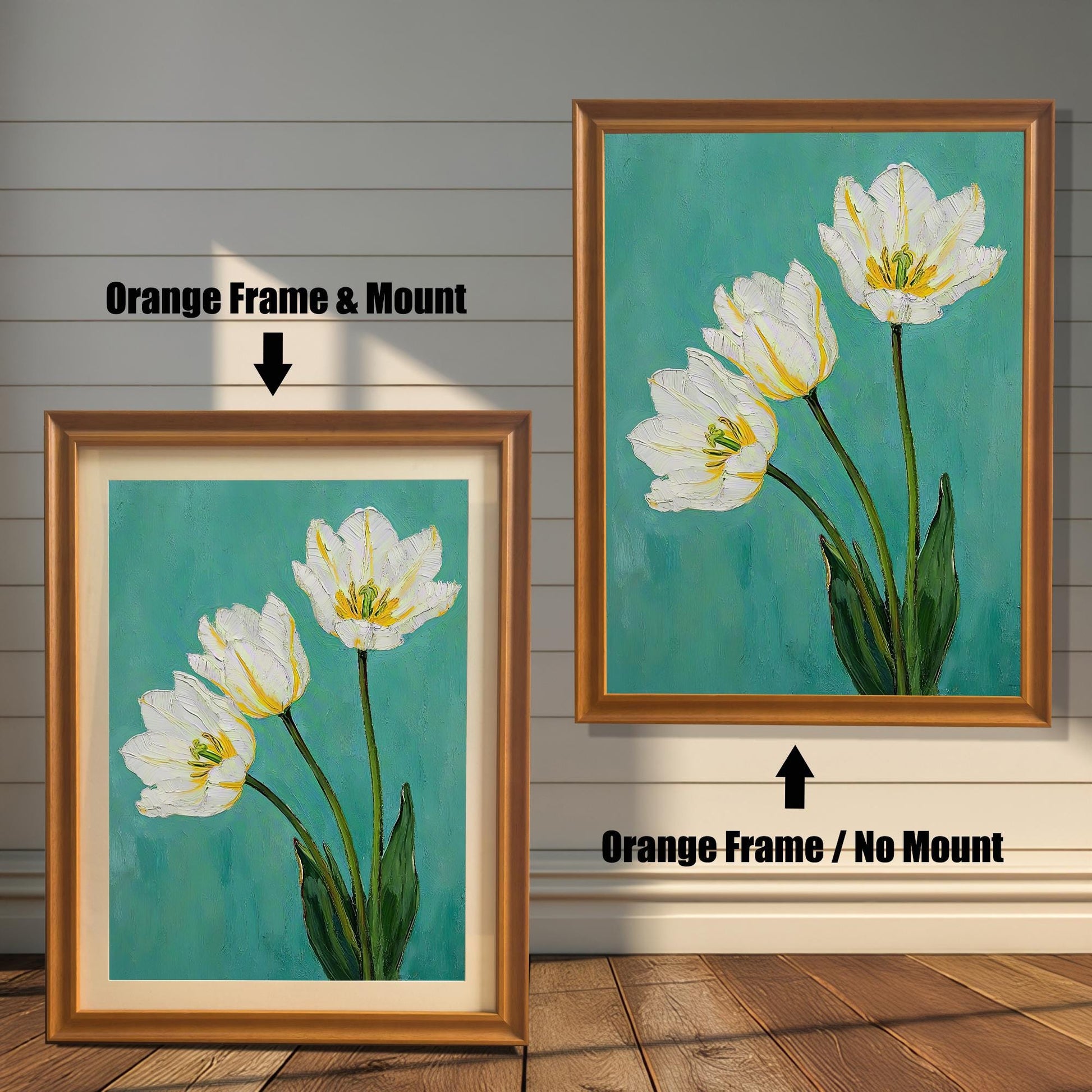 A framed art print of three white tulips. One is in a light orange-brown frame with a tan mat, and the other is in the same frame without a mat. The art is against a white wooden wall.