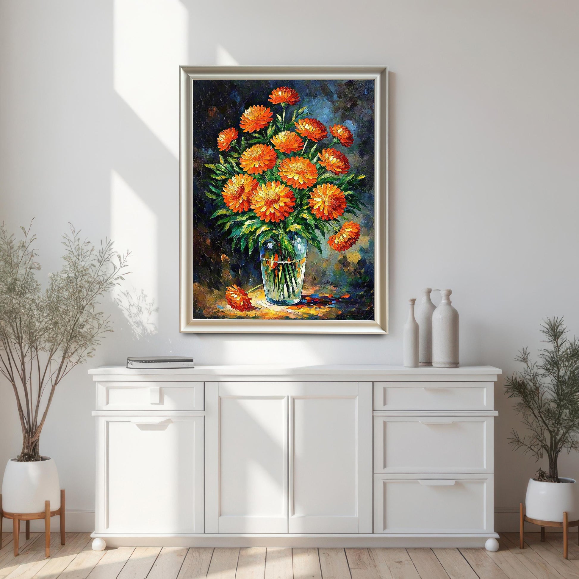 A white-framed art print of orange flowers hangs on a white wall above a white cabinet. Potted plants flank the cabinet.