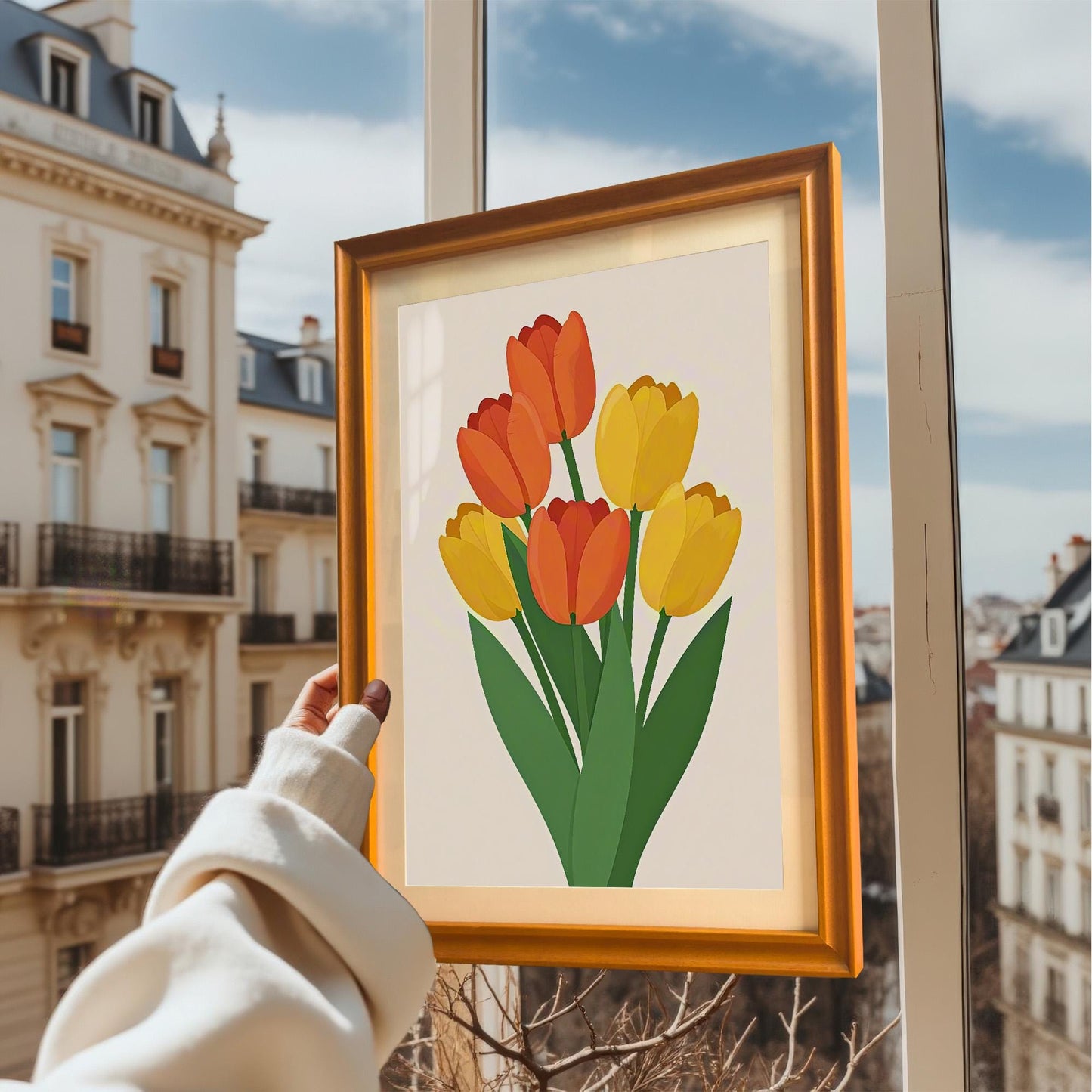 A hand holds up a framed art print of orange and yellow tulips against the backdrop of a building and sky.
