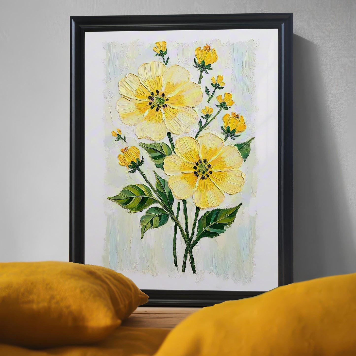 A close-up of a black-framed art print of yellow flowers with a textured, painterly style. Two blurred, yellow pillows are in the foreground.
