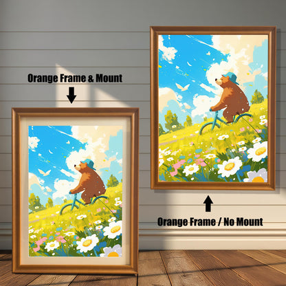 A split image showing the art print in two orange-toned wood frame options. The left print has an orange frame with a white mat, and the right print is in an orange frame without a mat. The cheerful art features a brown bear in a blue hat riding a bicycle through a sunny field of flowers.