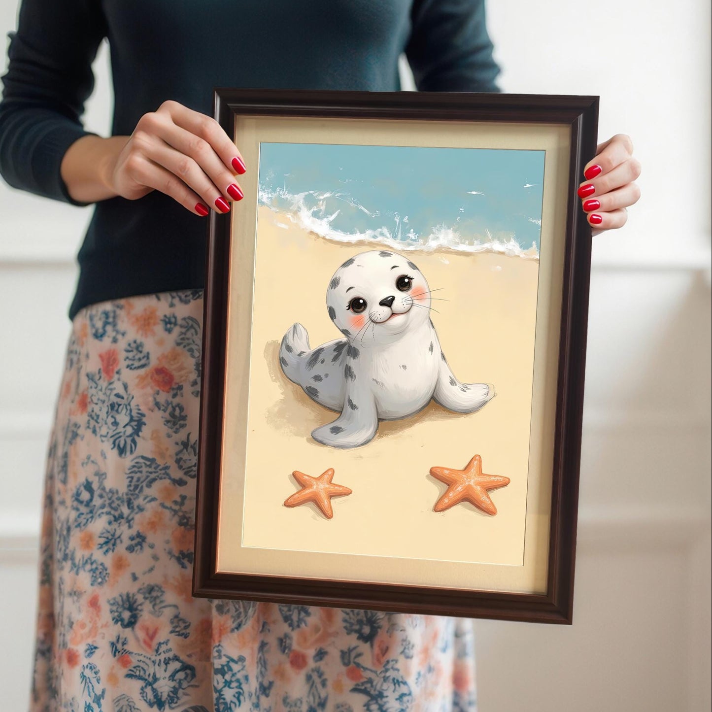 A person with red-painted fingernails holds a framed wall art print of a cute baby seal on a beach. The print is in a coffee-colored frame with a mount. The person's face is not visible.