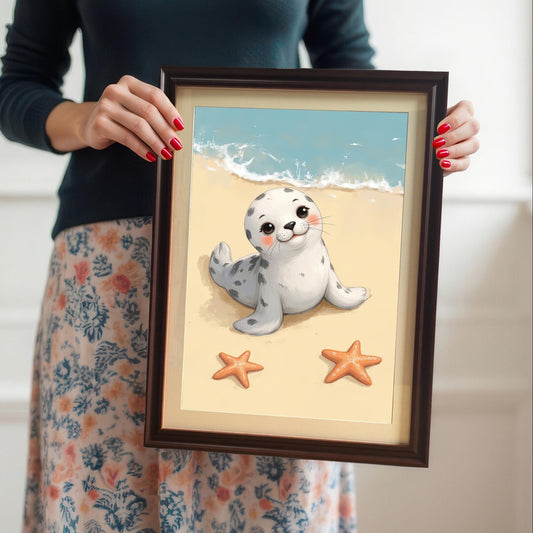 A person with red-painted fingernails holds a framed wall art print of a cute baby seal on a beach. The print is in a coffee-colored frame with a mount. The person's face is not visible.