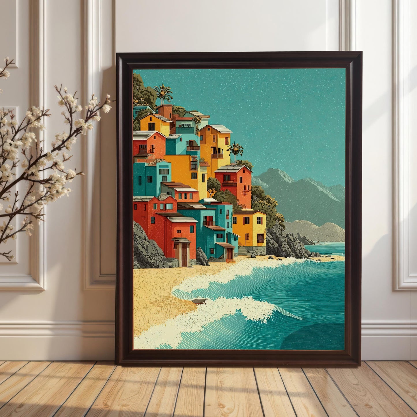 A framed art print leans against a white wall with wainscoting. The print depicts colorful houses on a hill overlooking a beach and ocean. A vase with white flowers sits to the left.