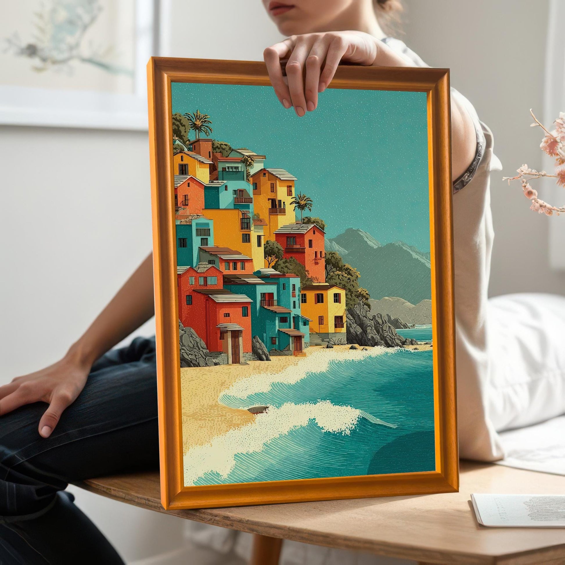A person sitting on a table holds up a framed art print. The print shows a collection of colorful houses on a sandy beach next to a turquoise ocean.