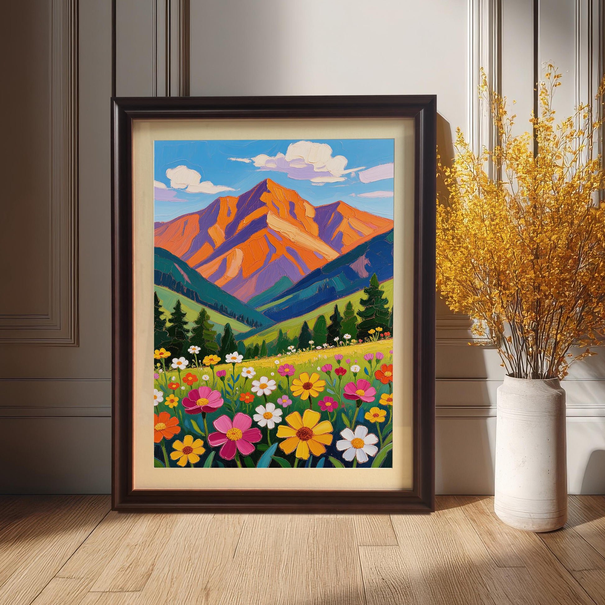 A dark-framed painting of a vibrant mountain scene with a field of wildflowers leans against a gray paneled wall next to a vase with dried yellow flowers.