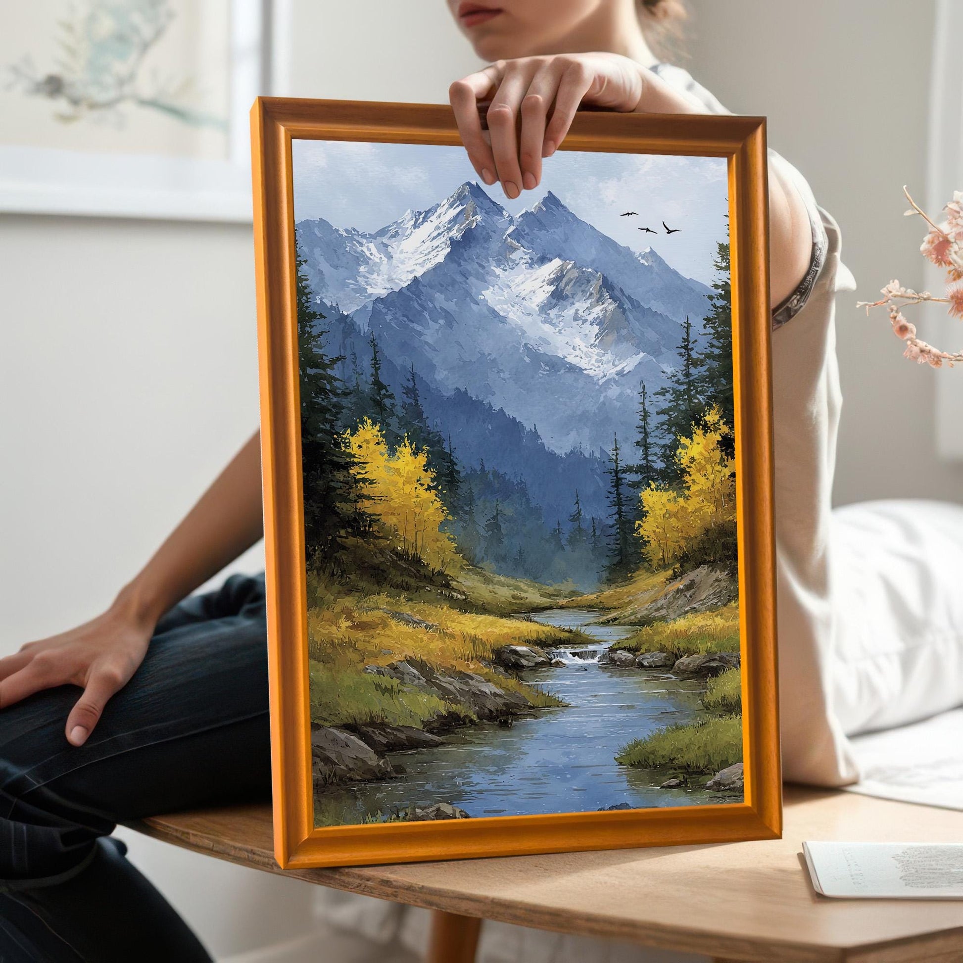 A person holds a gold-framed art print of a mountain landscape with a stream and yellow trees.