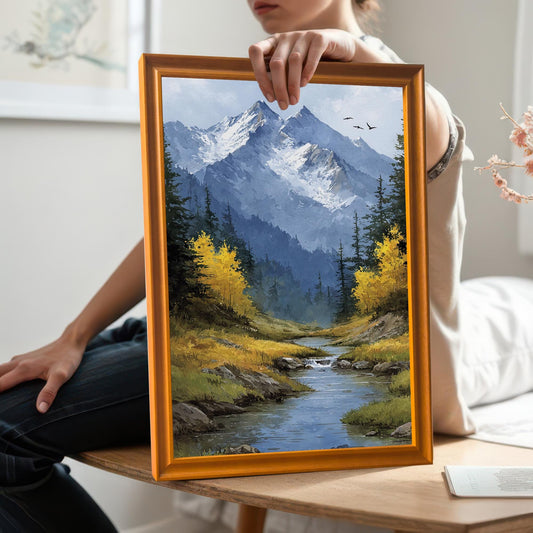 A person holds a gold-framed art print of a mountain landscape with a stream and yellow trees.