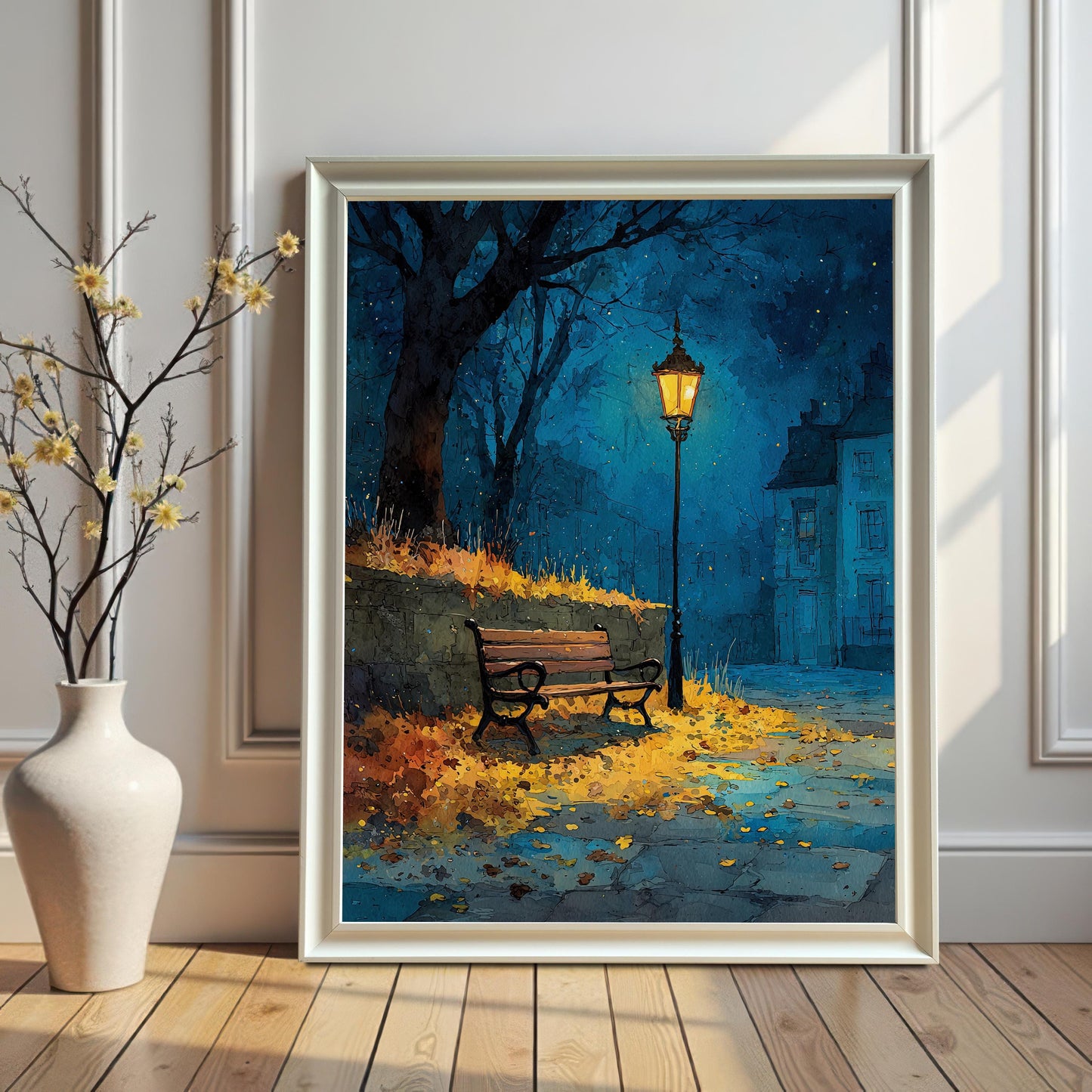 A framed painting of a street scene with a lamppost and a bench. The painting is in a white frame and leans against a white wall next to a vase with yellow flowers.