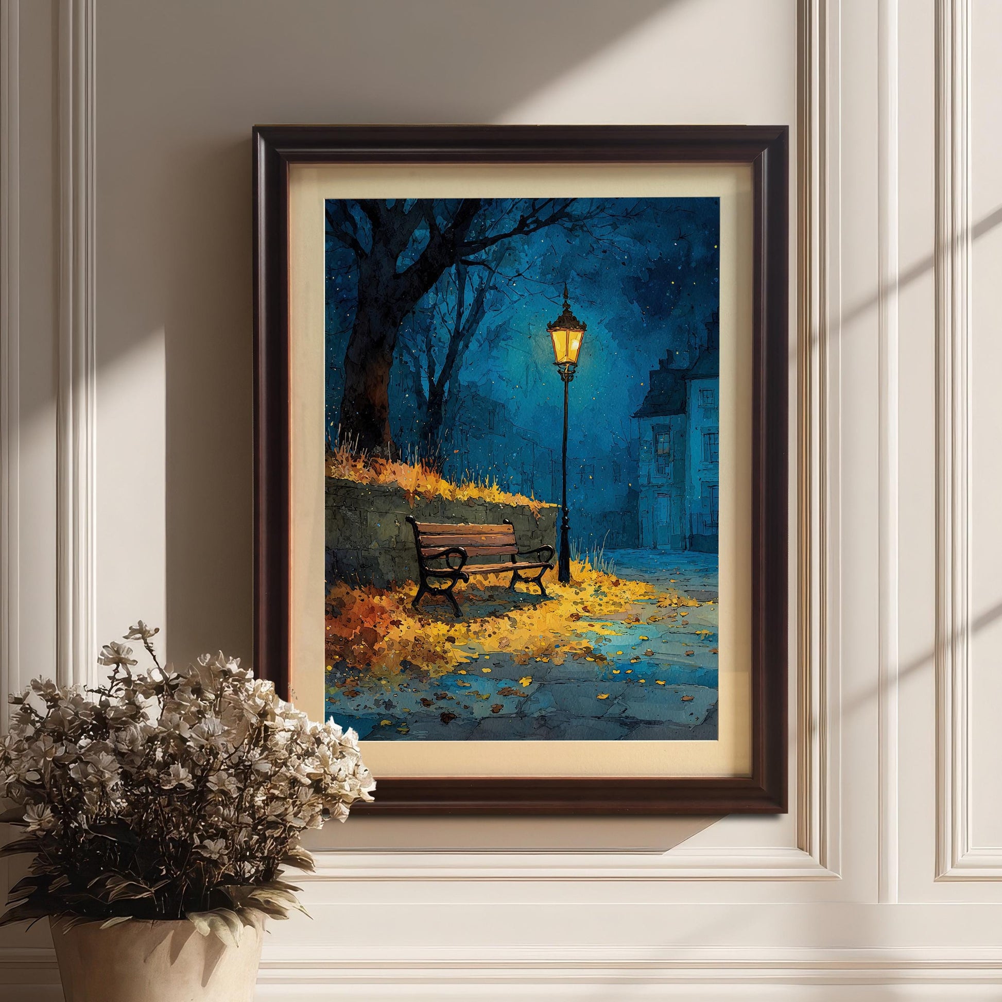 A framed painting of a street with a lamppost and a bench. The painting is in a coffee-colored frame and is leaning against a white wall next to a pot of white flowers.