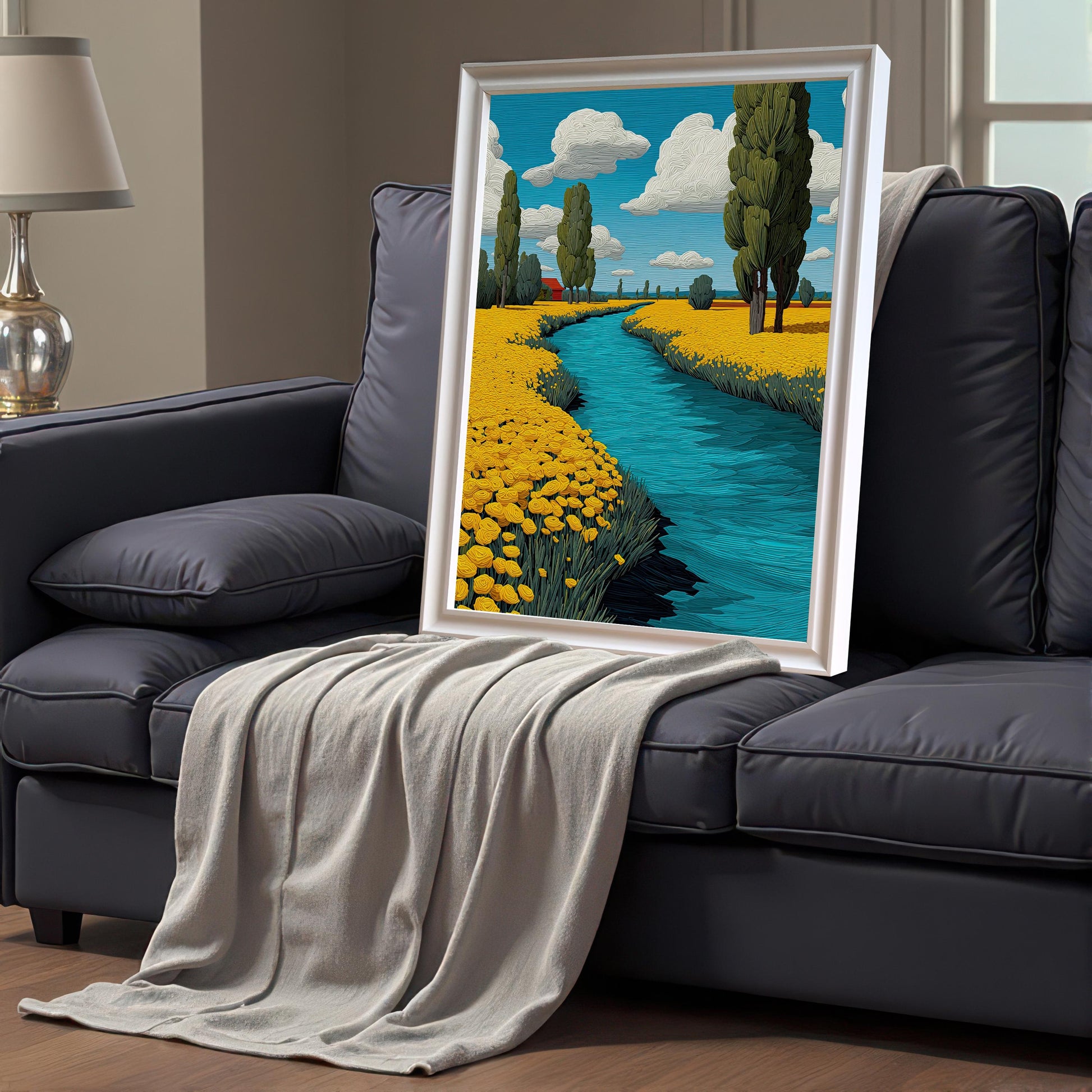 A white-framed painting of a river flowing through a field of yellow flowers. The painting is leaning against a dark gray couch with a blanket draped over it.