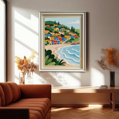 A framed painting of a village with colorful houses on a hill overlooking a beach. The painting, in a shiny silver frame, is hanging on a wall above a wooden table.