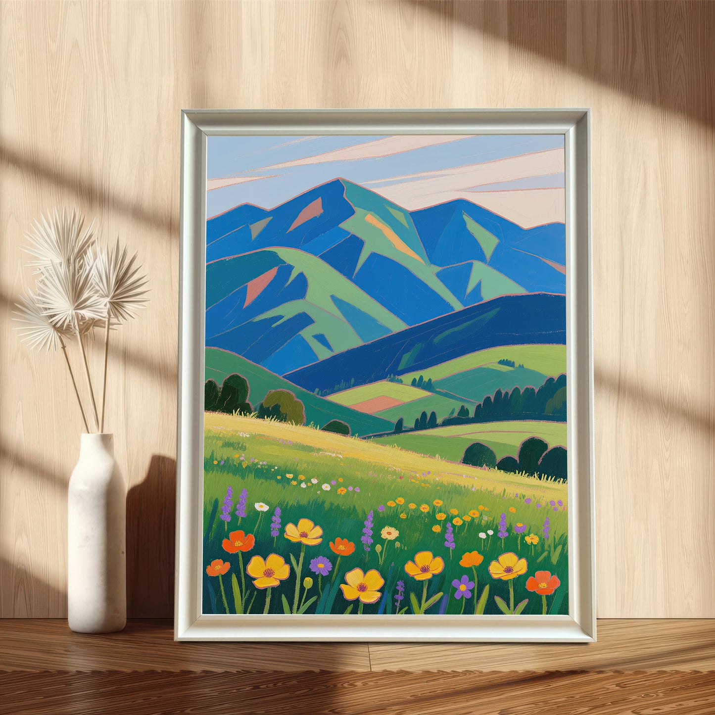A framed painting of a meadow with colorful flowers and mountains in the background. The painting is in a white frame and is leaning against a wooden wall next to a vase.
