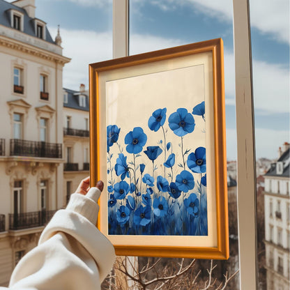 A person holds up a framed print of blue flowers against a window, showing the art with a European cityscape in the background.