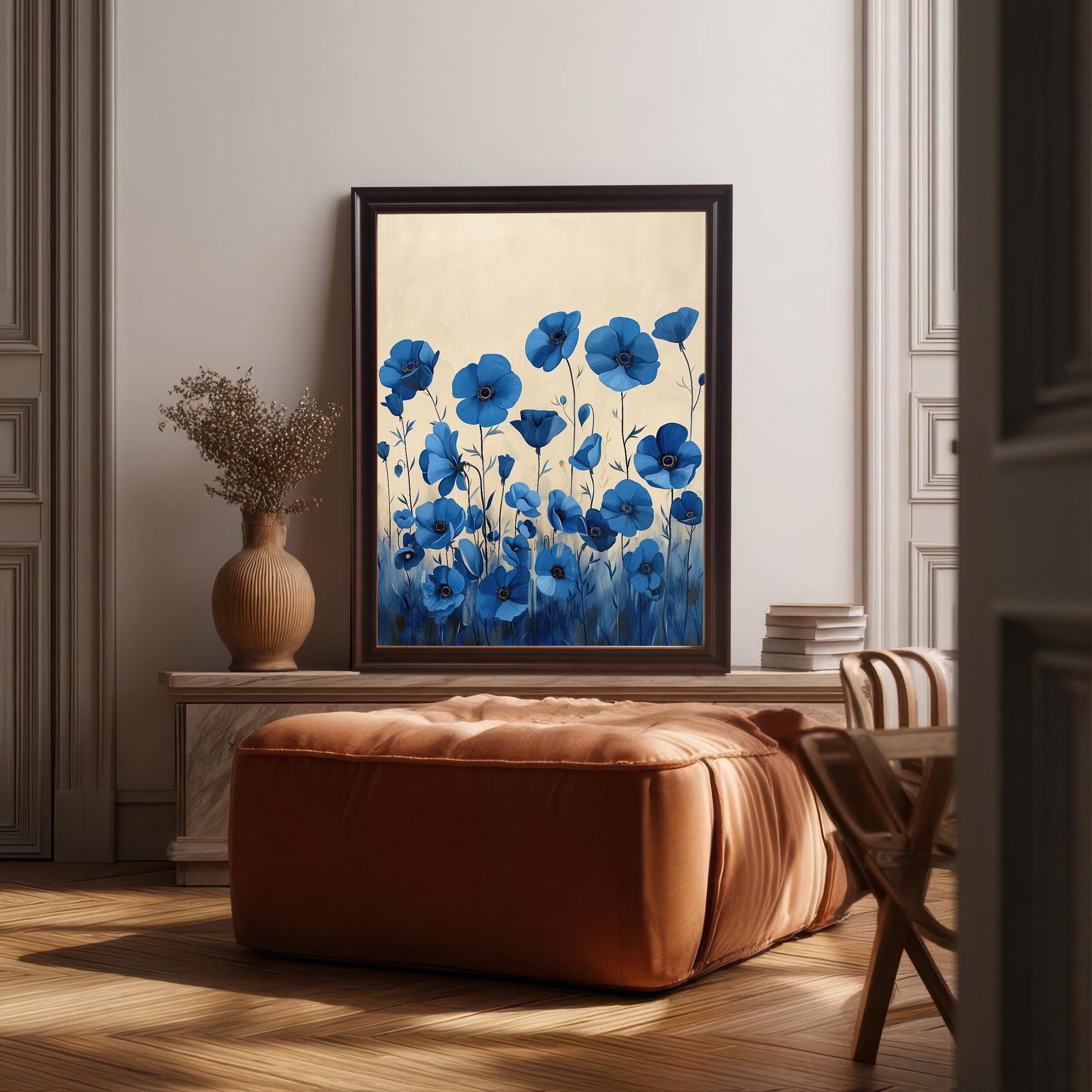 A framed art print of blue flowers with a light background. The dark brown-framed art rests on a wooden chest behind a brown ottoman and a vase with dried flowers.