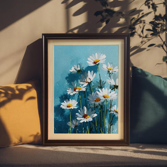 A framed art print of white daisies on a blue background, with a dark wood frame, resting on a couch between two pillows.