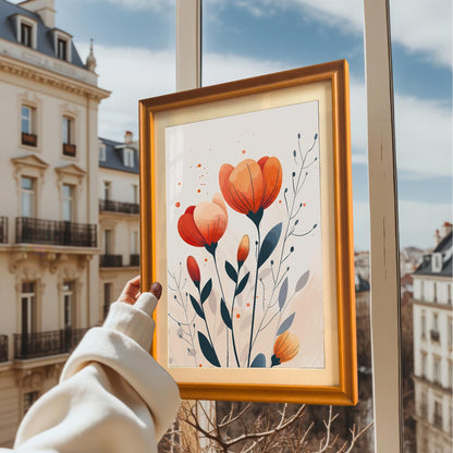 A person holds up a framed print of orange and red flowers against a window, showing the art with a European cityscape in the background.