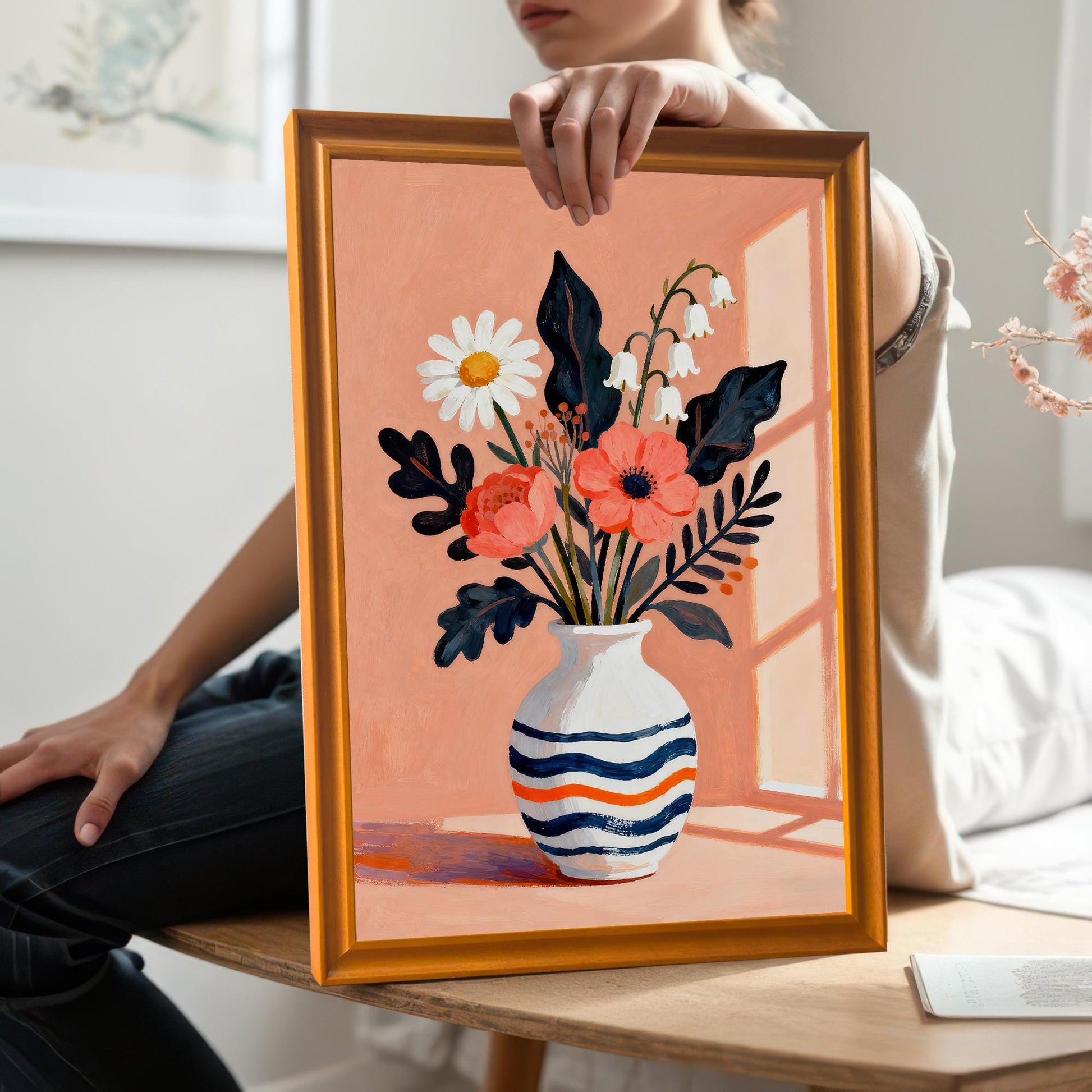 A person holding a framed floral art print in an orange frame. The print is angled towards the camera, showing a close-up of the artwork.