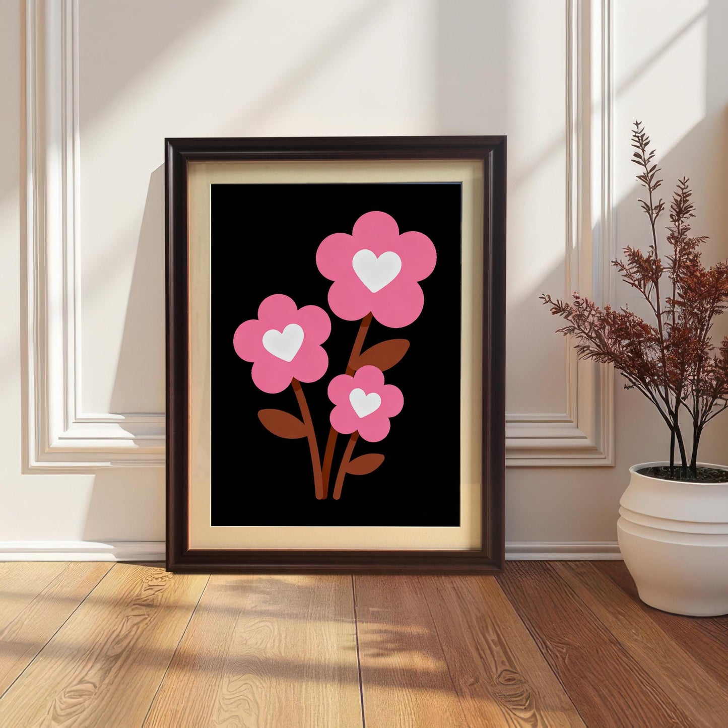 A framed art print of three pink flowers with white hearts leans against a white paneled wall. The frame is a dark brown color, and a houseplant sits next to it.
