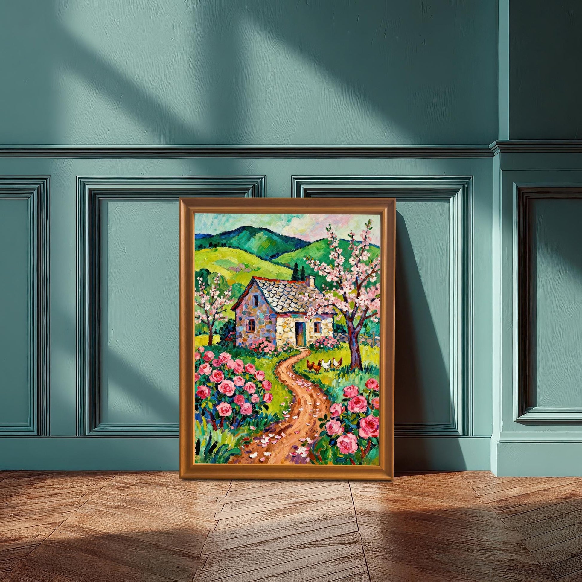 A framed, textured oil painting of a cottage with a rose garden. The frame is propped on a wooden floor against a paneled teal wall with sunlight shadows.