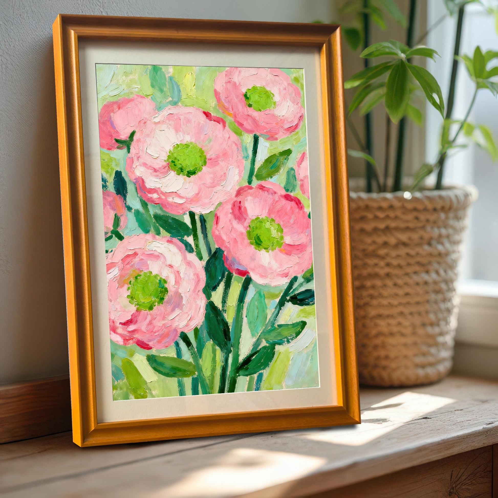 A painting of pink flowers in a gold frame propped on a wooden windowsill next to a houseplant in a woven pot.