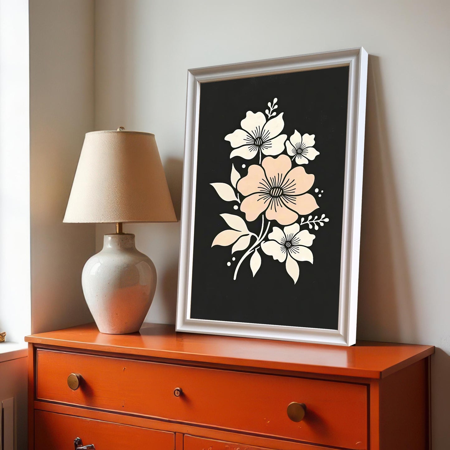 A framed floral art print featuring a central pink flower surrounded by white blooms on a dark background. It is propped on a bright orange dresser next to a lamp.