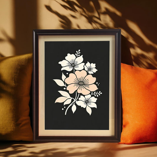 A framed floral art print featuring a central pink flower surrounded by white blooms on a dark background. It is propped on a surface between two pillows, with shadows of leaves cast over it.