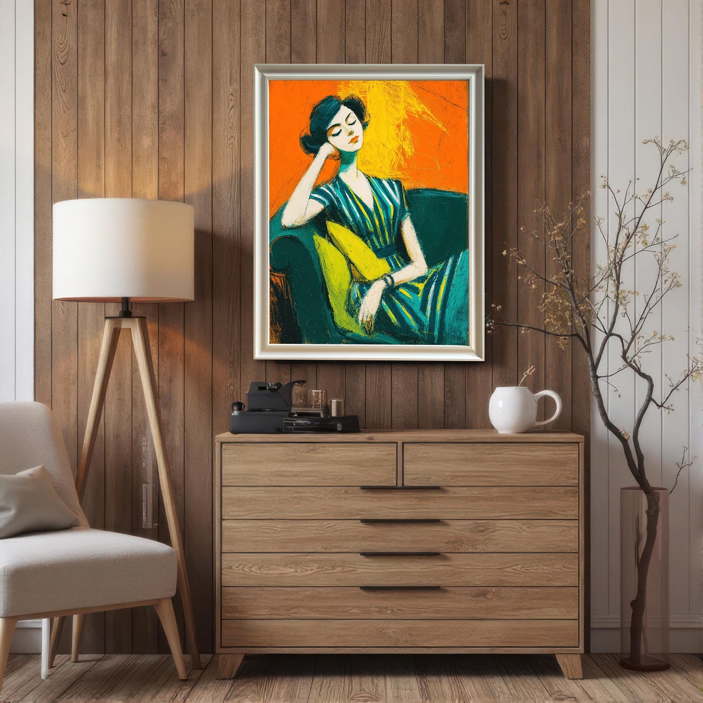 A framed art print of a seated woman in a silver frame, hanging on a wood-paneled wall above a light wood dresser. A floor lamp and plant are on either side.