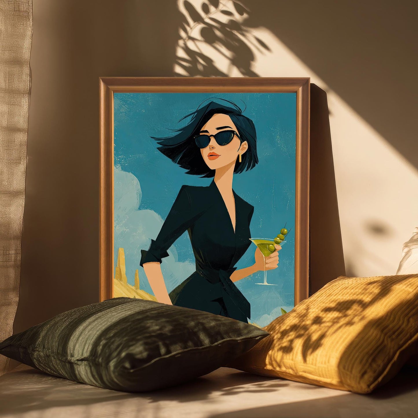 A framed art print of a woman in a black dress holding a martini. The orange-framed print is propped on a couch between a black and a yellow pillow.