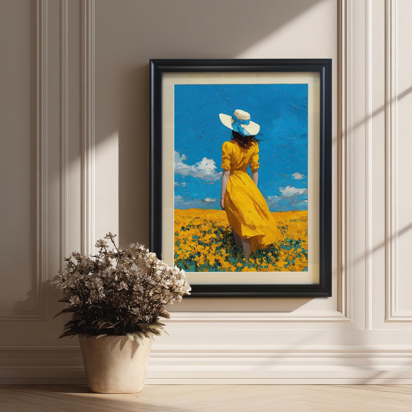 A black-framed art print of a woman in a yellow dress in a field, hanging on a white paneled wall with a plant on a side table.