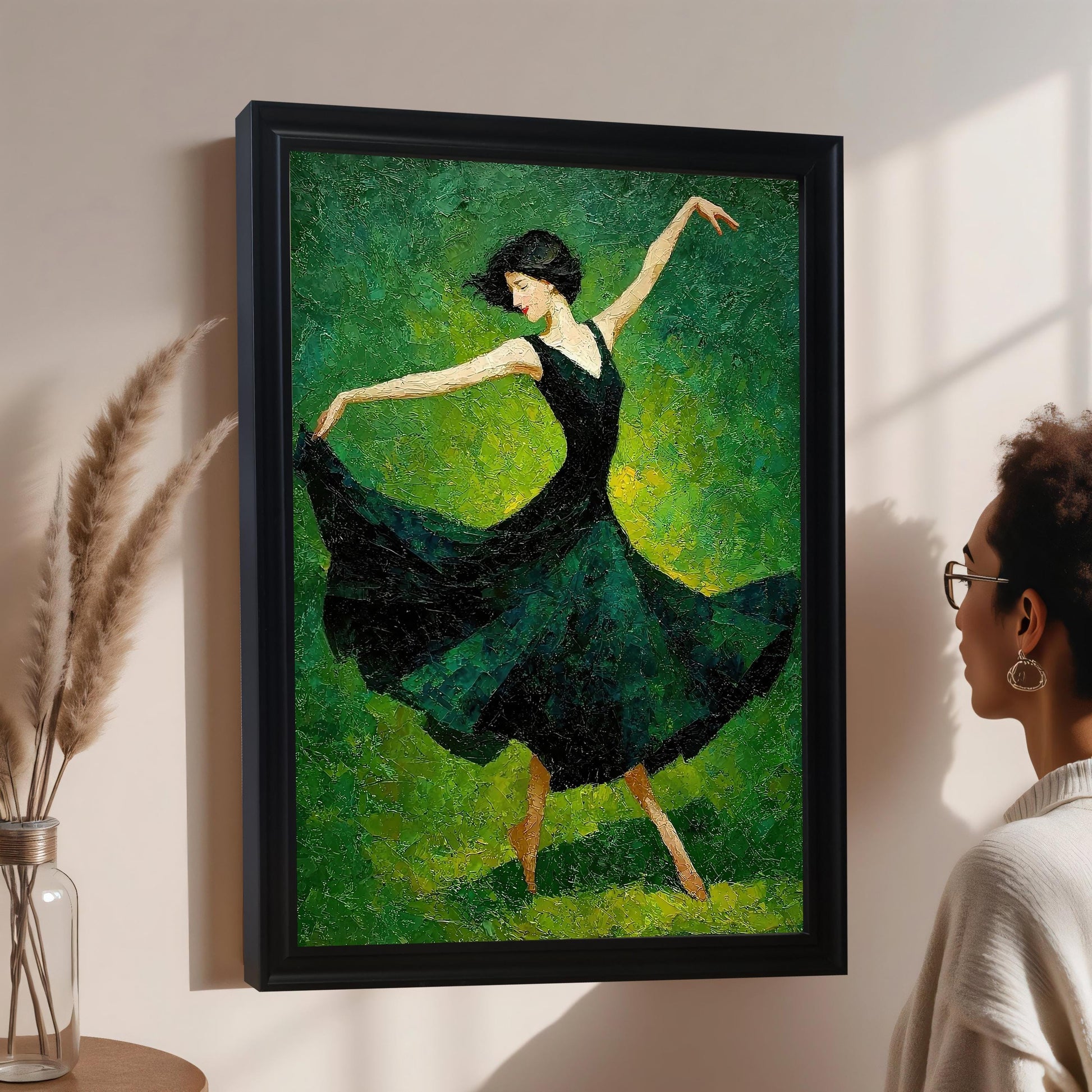 A woman with glasses looks at a framed painting of a dancer in a dark green dress. The painting hangs on a light pink wall next to a glass vase with dried reeds.